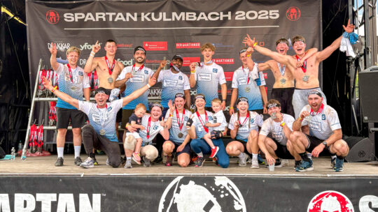 CONFIDO nach dem Spartan Race 2025 in Kulmbach – alle gemeinsam durch das Ziel. Das Bild zeigt das gesamte Team auf der Bühne – mit Kindern und Familie. Auch die, die nicht mitgelaufen sind, sind dabei.