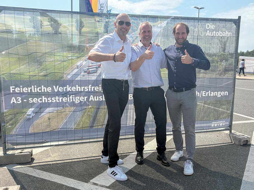 CONFIDO Ingenieure stehen mit Daumen hoch in der Sonne vor dem Bauzaunbanner von "Die Autobahn"