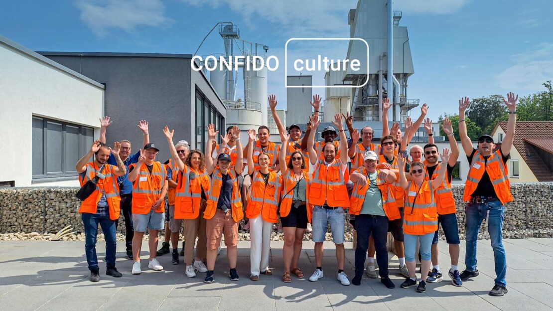 Confido Ingenieure auf Werksbesichtigung: vor dem Betonwerk in der Sonne werfen sie ihre Arme in die Luft