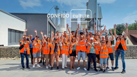 Confido Ingenieure auf Werksbesichtigung: vor dem Betonwerk in der Sonne werfen sie ihre Arme in die Luft