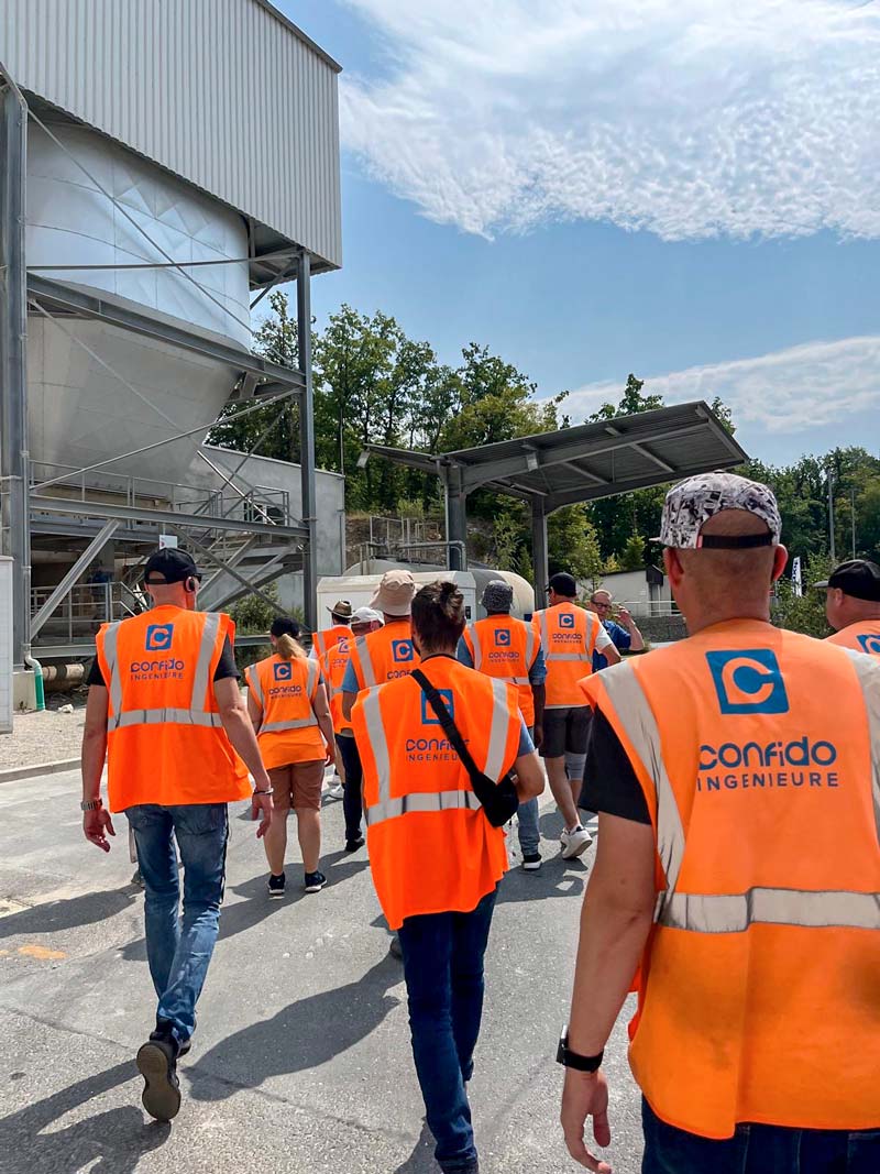 Confido Ingenieure von hinten auf dem Weg durch das Betonwerk: alle tragen Signal-Orangene Warnwesten mit dem Unternehmenslogo bei herrlichem Sonnenschein.